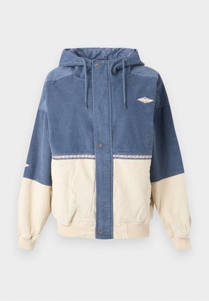 Μπλε και μπεζ μπουφάν από κεντητό κοτλέ με κουκούλα, κουμπιά σούστες μπροστά, κορδόνια, δύο τσέπες και μικρά κεντημένα μπαλώματα στο στήθος και στο μανίκι.