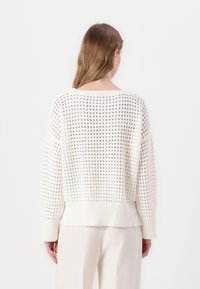 Woman with shoulder-length light brown hair wearing a loose, white, open-knit sweater and cream trousers, shown from the back on a white background.