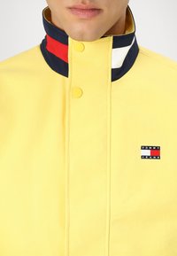 Gelbe Jacke mit hohem Kragen, ausgestattet mit marineblauen, roten und weißen Akzenten. Kleines Tommy Jeans-Logo auf der linken Brust. Glatte Textur.