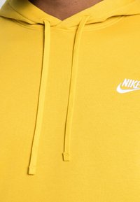Žlutá mikina s kapucí z měkké tkaniny má stahovací šňůrku a bílé logo Nike na levé straně hrudníku, poskytující hladkou texturu.
