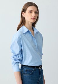 Lichtblauw gestreept shirt met knoopsluiting, brede kraag en opgerolde mouwen, gecombineerd met donkerblauwe jeans en een zwarte riem.