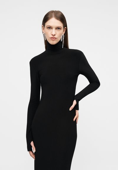 Γυναίκα που φοράει εφαρμοστό μαύρο φόρεμα ζιβάγκο με μακριά μανίκια και κρεμαστά σκουλαρίκια με κρύσταλλα, ποζάροντας με το ένα χέρι στη μέση.