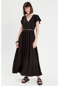 Abito maxi nero con maniche corte a balze e scollatura a V. Presenta una vita aderente con dettagli in pizzo, gonna fluida e sandali piatti.