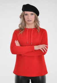 Rode sweatshirt met waffle-textuur, voorzien van een capuchon en zilverkleurige trekkoorden. Draagt met zwarte leggings en een zwarte baret.