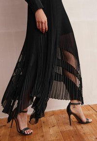 Robe plissée noire avec des accents transparents, design superposé et silhouette fluide. Portée avec des sandales à talons hauts noires à lanières sur un sol en bois.