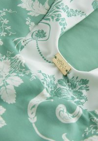 Žalias mėtų spalvos raštuotas lengvas audinys su baltais gėlių motyvais. Yra auksinės spalvos žymė, iškilmingai graviruota gėlių motyvu.