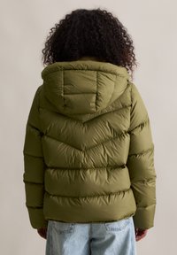 Oliwkowa puchówka z kapturem, z chevronowym przeszyciem i pikowanym wzorem. Sylwetka jest skrócona, w połączeniu z jasnoniebieskimi dżinsami.