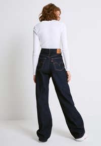 Jean en denim foncé taille haute avec jambes larges, orné de coutures contrastantes et deux poches arrière, associé à un haut blanc ajusté à manches longues.