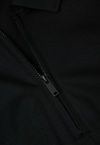 Camisa polo negra con cierre de media cremallera, que presenta una textura suave y un tirador de cremallera en contraste, sobre un fondo negro liso.