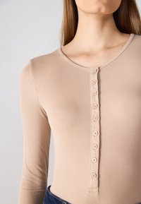 Body à manches longues, côtelé en beige avec un col rond et une rangée de sept petits boutons au centre. Texture douce, design ajusté.