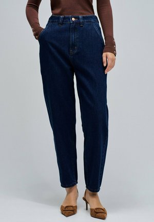 Mujer con jeans de tiro alto ajustados de color azul oscuro, una blusa de manga larga marrón con botones en las mangas, y zapatos de tacón marrones con punta.