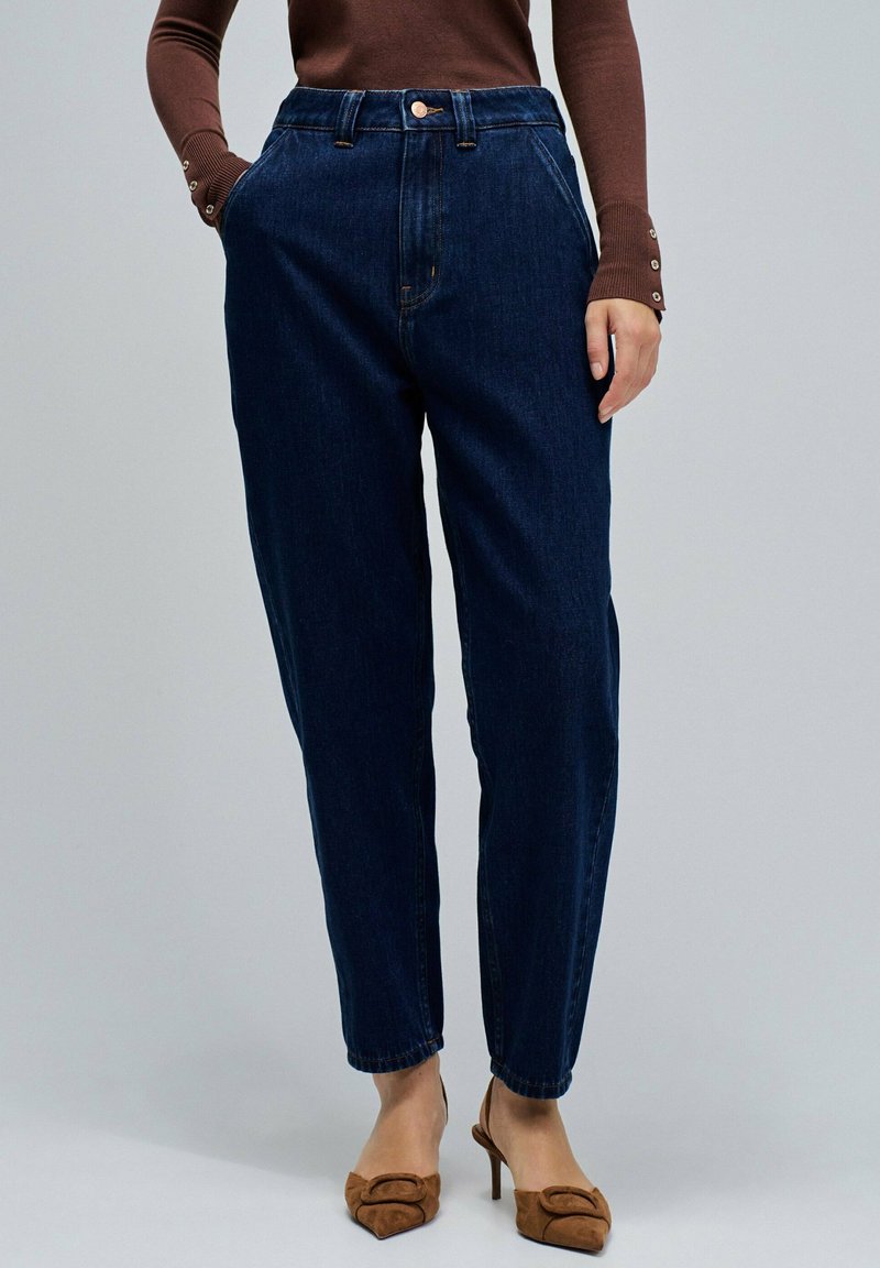 Donna che indossa jeans a vita alta di colore blu scuro, un top lungo marrone con bottoni sulle maniche e scarpe con tacco a punta marroni.
