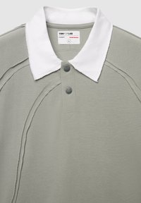 Polo grigio con colletto bianco, dotato di tre bottoni neri, tessuto testurizzato e cuciture rifinite lungo le cuciture.