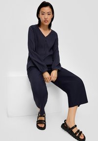 Dunkelblusige langärmelige Bluse mit V-Ausschnitt, die eine subtile Textur und Knopfdetails aufweist, kombiniert mit weiten navyblauen Hosen und schwarzen Sandalen.