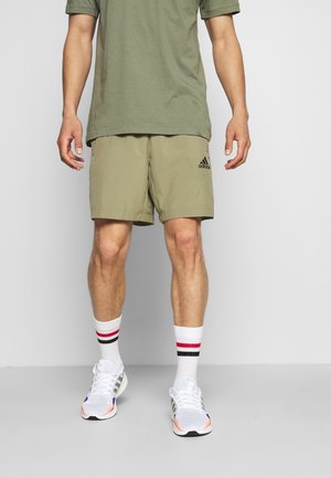 Man draagt olijfgroene Adidas short, groen T-shirt, witte sportsokken met rode en zwarte strepen, en witte hardloopschoenen, staand tegen een grijze achtergrond.