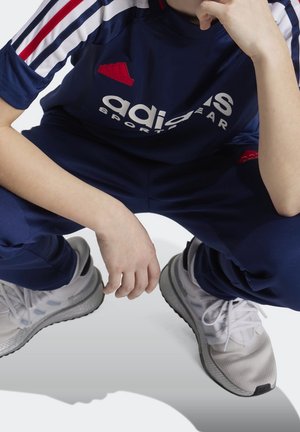 Conjunto de ropa deportiva Adidas en azul marino con rayas rojas y blancas. La camiseta presenta un logo blanco, combinada con pantalones deportivos azul marino y zapatillas gris claro.