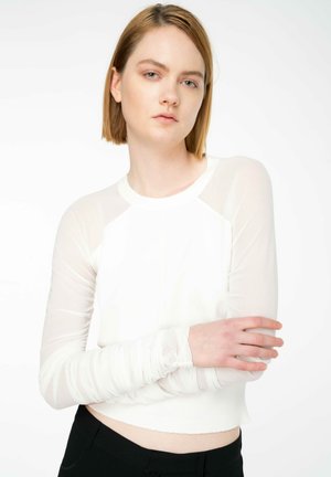 Top lungo bianco a maniche lunghe con un corpo aderente, maniche trasparenti e polsi arricciati. Progettato con un collo rotondo e un orlo corto.