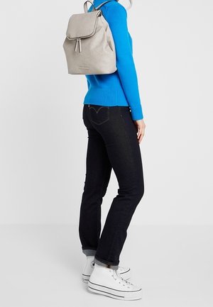 Personne portant un pull bleu vif, un jean foncé, des baskets blanches montantes et un sac à dos beige clair, debout devant un fond uni.