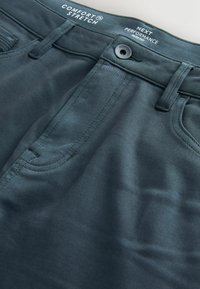 Kényelmes, stretch anyagú nadrág sötét türkiz színben, sima textúrával, gombos záródással és diszkrét varrási részletekkel. Elöl zsebekkel rendelkezik.