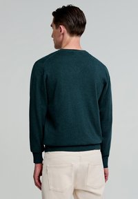 Pulover verde închis, cu guler rotund, cu mâneci lungi, cu manșete și tiv ribbate. Textură moale, design simplu, purtat cu pantaloni de culoare deschisă.