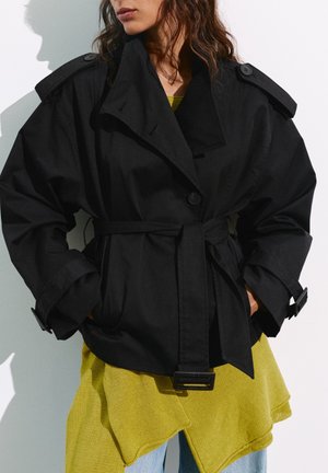 Femme portant une veste noire oversize ceinturée sur un top jaune à ourlet asymétrique et un jean bleu clair, mains dans les poches, légèrement tournée vers la gauche.
