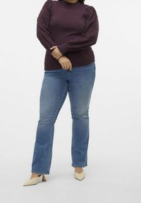 Donna che indossa un maglione viola scuro con maniche a sbuffo testurizzate, jeans blu a gamba dritta e décolleté crema a punta, con le braccia incrociate.