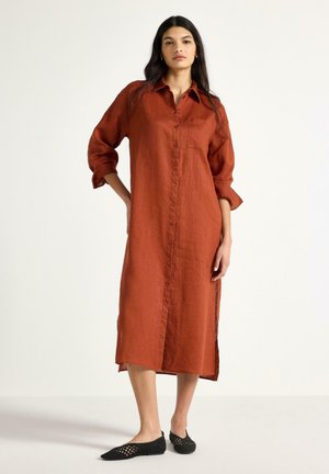 Kvinde med langt mørkt hår iført en rustrød midi skjortekjole med knapper og op rullede ærmer samt sorte flettede flade sko, stående mod en hvid baggrund.
