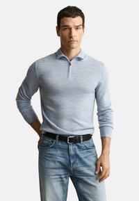 Mann in hellblauem langärmligen Reißverschluss-Pullover, der in verblasste blaue Jeans mit schwarzem Gürtel gesteckt ist, steht vor einem schlichten Hintergrund.