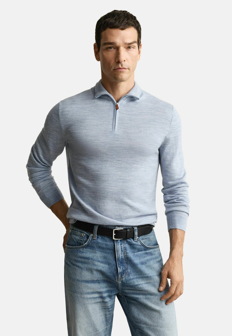 Mann in hellblauem langärmligen Reißverschluss-Pullover, der in verblasste blaue Jeans mit schwarzem Gürtel gesteckt ist, steht vor einem schlichten Hintergrund.