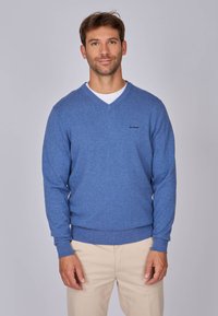 Pull en v bleu en tissu doux, avec une texture tricotée et des poignets côtelés. Porté par-dessus une chemise blanche avec un logo discret sur la poitrine.