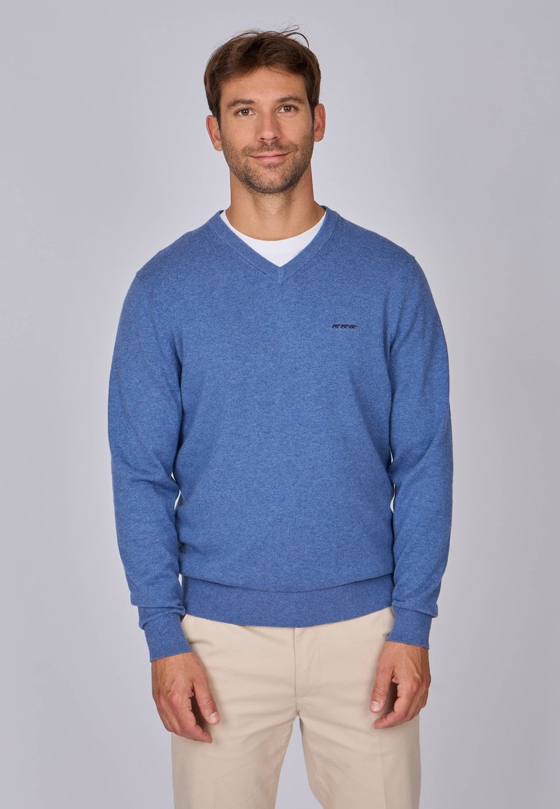 Pull en v bleu en tissu doux, avec une texture tricotée et des poignets côtelés. Porté par-dessus une chemise blanche avec un logo discret sur la poitrine.