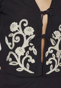 Vestuário preto com um decote em V profundo, apresentando intrincados bordados florais brancos e um fecho com laço no decote. Textura de tecido suave.