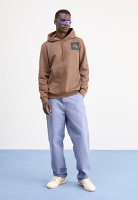 Bruine hoodie met een buidelzak en logo, gecombineerd met lichtblauwe, loszittende broek en witte sneakers. Model draagt blauwe zonnebril.