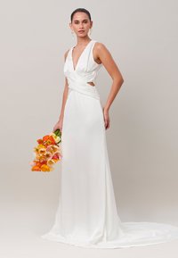 Femme en robe de mariée blanche sans manches avec un décolleté en V profond et des découpes latérales, tenant un bouquet de fleurs orange et jaunes.