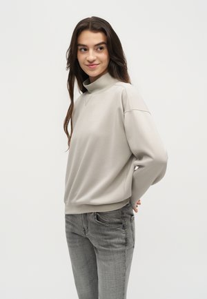 Jeune femme aux longs cheveux bruns porte un sweat à col roulé beige clair et un jean gris, légèrement de profil, les mains derrière le dos.