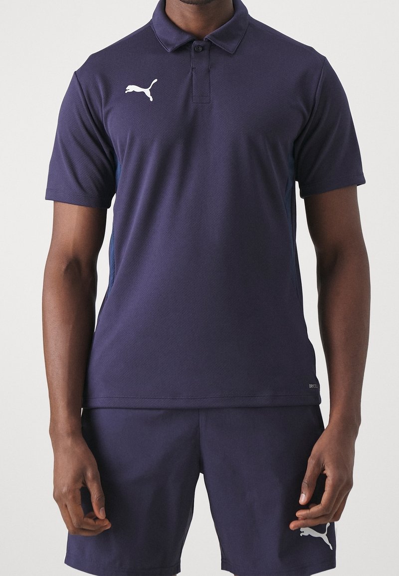 Homme portant un polo Puma bleu foncé et un short assorti, debout devant un fond clair uni.