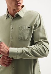 Lange mouwen, lichtgroen shirt van katoen. Heeft een button-down kraag, een borstzak en bruine knopen. Zachte textuur met een relaxte pasvorm.