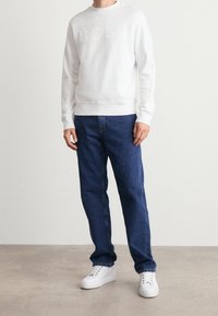 Vit sweatshirt med "BOSS"-logotyp i mjuk textur, tillsammans med blå denimjeans och vita sneakers, vilket ger en avslappnad passform.
