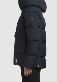 Granatowa pikowana kurtka z kapturem, o fakturowanej powierzchni, dużymi przednimi kieszeniami i okrągłą naszywką z logo na rękawie.