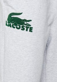 Grå bomullsstoff med en grønn brodert krokodillelogo og teksten "LACOSTE" under i fet skrift. Enkelt design, uformell stil.