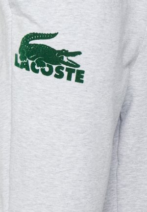 Szara bawełniana tkanina z zielonym haftowanym logo krokodyla oraz tekstem "LACOSTE" poniżej w pogrubionej czcionce. Prosty design, casualowy styl.