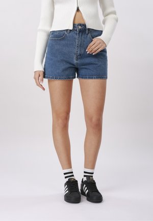 Femme portant un short en jean bleu, un pull zippé blanc côtelé, des baskets Adidas noires et des chaussettes blanches à rayures noires, debout devant un fond blanc.