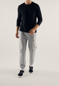 Černé žebrované tričko s dlouhým rukávem v kombinaci se světle šedými tepláky s dvěma velkými předními kapsami a zakončené manžetou. Černé tenisky.