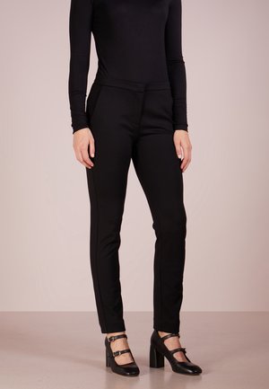 Person trägt eng anliegende schwarze Hose, schwarzes langärmeliges Oberteil und schwarze Mary Jane Absätze und steht auf einem hellen Boden vor einem einfarbigen Hintergrund.