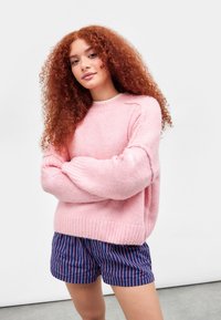 Rosa, flauschiger Pullover mit langen Ärmeln und geripptem Saum, kombiniert mit navy-blauen gestreiften Shorts. Vor einem schlichten weißen Hintergrund.