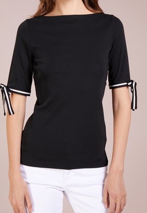 Femme portant un haut noir ajusté à manches mi-longues avec des nœuds en ruban rayé noir et blanc, assorti à un pantalon blanc.