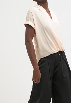 Femme portant un chemisier cache-cœur à manches courtes beige clair, rentré dans un pantalon ample noir taille haute avec une ceinture à boucle argentée.