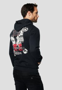 Czarna bluza z kapturem z nadrukiem graficznym z tyłu, zawierającym tekst i ilustracje. Miękki materiał, luźny krój, ściągacze na mankietach i u dołu. Noszona także z ciemnymi jeansami.