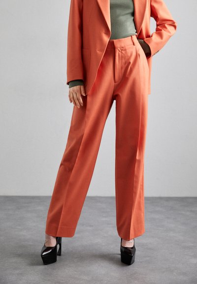 Oranžový ušitý blazer a ladící kalhoty s vysokým pasem z hladké látky; doplněno o přiléhavý zelený top a černé platformové podpatky.