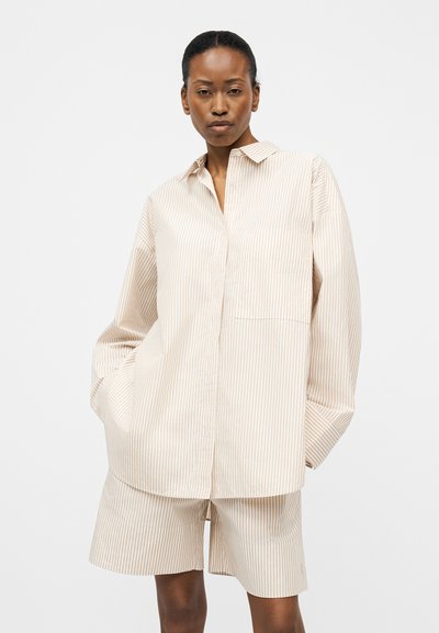 Γυναίκα που φοράει μπεζ και λευκό πουκάμισο με κάθετες ρίγες σε φαρδιά γραμμή και ταιριαστά σορτς μέχρι το γόνατο, στέκεται με τα χέρια στις τσέπες.